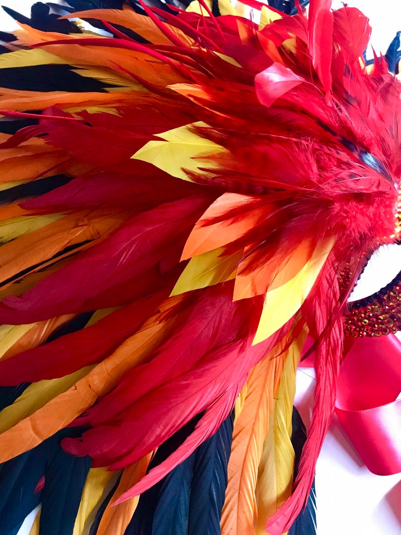May include: A vibrant headdress featuring a cascade of red, orange, yellow, and black feathers. The feathers are arranged in a layered pattern, creating a dynamic and eye-catching design. A red ribbon and jeweled accents are also visible.