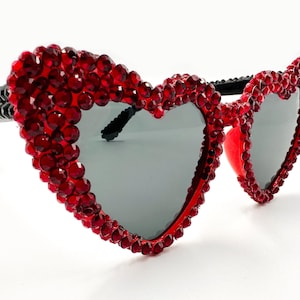 May include: Heart-shaped sunglasses with red jewel embellishments. The frames are black, and the lenses are gray. The sunglasses are set against a white background.