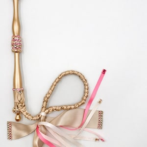 May include: A gold-coloured decorative wand with pink gemstone accents. The wand features a braided handle and is adorned with ribbons in shades of pink and cream. The ribbons are embellished with pink gemstones.