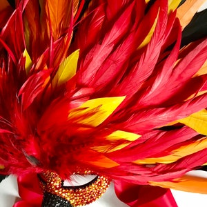 May include: A vibrant, decorative mask with a red, orange, and yellow feather design. The mask features a jeweled eye and beak, with a red satin ribbon. The mask is likely for a costume or masquerade.