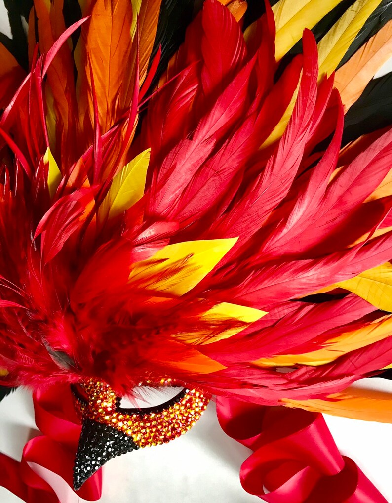 May include: A vibrant, decorative mask with a red, orange, and yellow feather design. The mask features a jeweled eye and beak, with a red satin ribbon. The mask is likely for a costume or masquerade.
