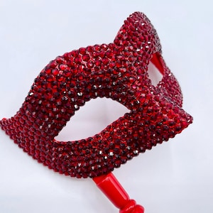 May include: A red, jewel-encrusted masquerade mask with a matching handle. The mask is covered in small, sparkling red gems, creating a dazzling effect. The eye openings are oval-shaped. The handle is a solid red color.