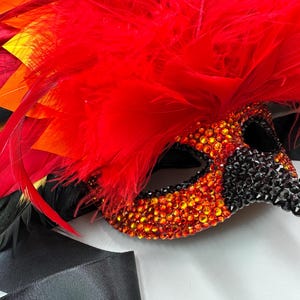 May include: A decorative masquerade mask with a red feather plume and black ribbon. The mask is embellished with orange and black rhinestones. The feathers are red, orange, and black.