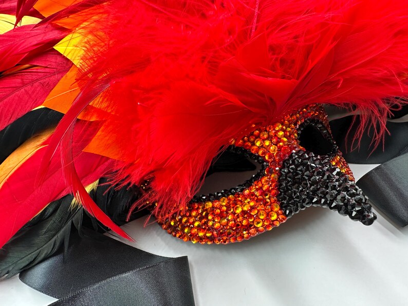 May include: A decorative masquerade mask with a red feather plume and black ribbon. The mask is embellished with orange and black rhinestones. The feathers are red, orange, and black.