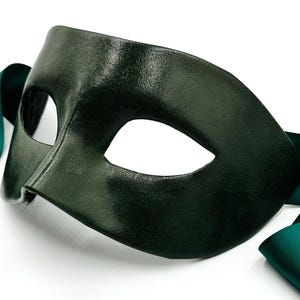 Könnte beinhalten: Eine dunkelwaldgrüne Maskenballmaske mit glatter, lederartiger Textur und zwei mandelförmigen Augenöffnungen. Die Maske verfügt über lange, passende dunkelgrüne Satinbänder, die an den Seiten befestigt sind, um sie am Kopf zu befestigen.