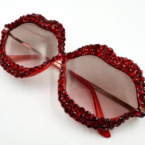 May include: Red sunglasses with a unique, embellished design. The frames are covered in small, sparkling red gems. The lenses are a gradient tint, transitioning from a darker shade at the top to a lighter shade at the bottom. The arms are a thin, gold color.