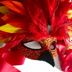 May include: A decorative mask with a phoenix theme. The mask is adorned with red, orange, and yellow feathers, and is embellished with sparkling orange and black rhinestones. A red satin ribbon is attached.