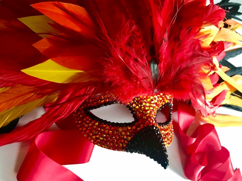 May include: A decorative mask with a phoenix theme. The mask is adorned with red, orange, and yellow feathers, and is embellished with sparkling orange and black rhinestones. A red satin ribbon is attached.