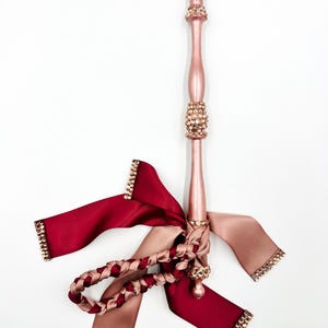 Könnte beinhalten: Ein dekoratives, rosa lackiertes Holzobjekt mit Kristallverzierungen. Angebracht sind bordeauxfarbene und beige Satinbänder, einige mit Kristallbordüren, und ein geflochtenes Band. Das Objekt befindet sich vor einem weißen Hintergrund.