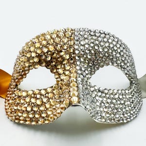 Peut inclure: Un masque décoratif divisé en deux parties, or et argent, incrusté de strass étincelants. Le masque comporte des ouvertures pour les yeux et est accompagné de rubans dorés et argentés, parfait pour un bal masqué.