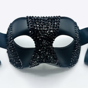 Pode incluir: Máscara preta de baile de máscaras com fita de cetim. A máscara é adornada com strass pretos nas bordas e no centro. A máscara tem um acabamento liso e fosco e é projetada para cobrir os olhos.
