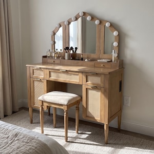 May include: A light brown wooden vanity table with a tri-fold mirror featuring integrated round light bulbs, cane-webbed cabinet doors, and a central drawer. A matching stool with a beige upholstered seat is positioned in front of the vanity.