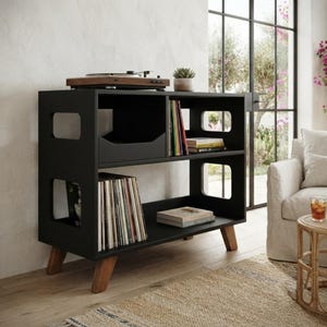 May include: A black wooden media console with two shelves, featuring cut-out side handles and angled light brown wooden legs. The top shelf holds a record player and books, while the bottom shelf contains a collection of vinyl records and books.