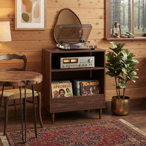 May include: A dark wood record player stand with two open shelves and a bottom drawer. The top shelf holds a silver audio receiver with two illuminated analog gauges. The bottom shelf contains vinyl records, including one titled Afrobeat.