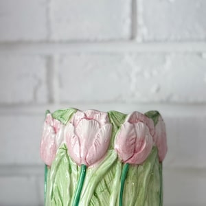 May include: A cylindrical ceramic planter featuring a three-dimensional relief design of light pink tulips with green stems and leaves. The surface has a glossy finish, and the sculpted floral pattern wraps around the entire exterior of the vessel.
