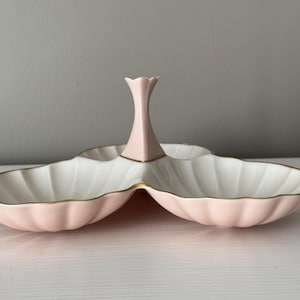 May include: A three-section serving dish with a central handle, featuring a scalloped shell-like design. The exterior is a soft pastel pink, while the interior is white, with delicate gold trim lining the edges of each compartment.