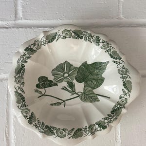 May include: A scalloped ceramic bowl featuring a cream-colored base with a dark green botanical transfer print. The center displays a morning glory flower and leaves, while the rim is decorated with a continuous vine of small flowers and foliage.