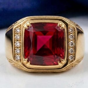May include: A gold ring featuring a large, square-cut, deep red gemstone set in the center. The band is accented with a vertical row of small, round, clear white stones on both sides of the central red gem, set within a polished gold frame.