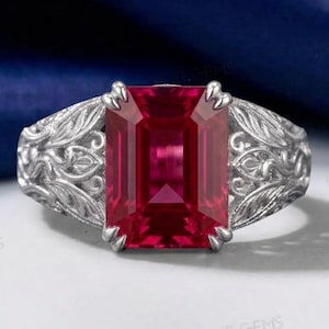 Può includere: Un anello color argento con una grande pietra preziosa rossa rettangolare a taglio smeraldo incastonata al centro. La fascia presenta intricati motivi in filigrana a rilievo a forma di foglia che si estendono dalla montatura centrale.