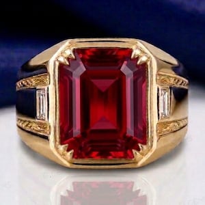 May include: A gold ring featuring a large, rectangular, emerald-cut red gemstone in the centre. The band is crafted from yellow gold and includes two small, clear, baguette-cut stones on either side, set within decorative, engraved gold metalwork.