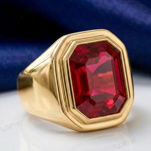 Può includere: Un anello in oro con una grande pietra preziosa rossa di taglio rettangolare a smeraldo incastonata in una fascia d'oro lucido. La montatura presenta una cornice geometrica a gradini che circonda la pietra rossa con sfaccettature.