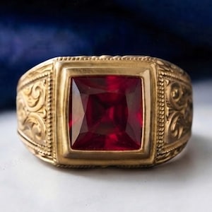 Könnte beinhalten: Ein goldener Ring mit einem quadratisch geschliffenen, tiefroten Edelstein in der Mitte. Die Ringschiene ist an den Seiten mit komplizierten, erhabenen Schnörkelmustern verziert, die für eine strukturierte Optik sorgen.
