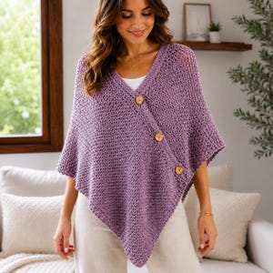 Puede incluir: Un poncho de punto color lavanda con cierre frontal asimétrico y tres botones redondos de madera marrón claro. La prenda presenta un escote en pico y un patrón texturizado de punto abierto en toda su superficie.