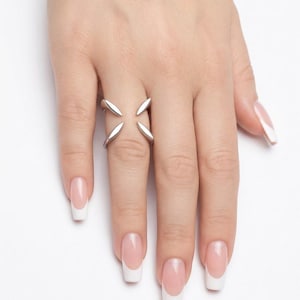 Peut inclure: Une bague argentée au design ouvert, composée de quatre éléments pointus en forme de pétales s'étendant vers l'extérieur. L'anneau est divisé, créant une disposition symétrique qui repose sur le majeur avec une finition polie et brillante.