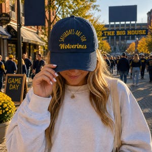May include: A navy blue baseball cap featuring the embroidered yellow text "SATURDAYS ARE FOR Wolverines" in a curved arch design across the front panels. The cap has a structured crown, a curved brim, and a matching navy blue button on the top.