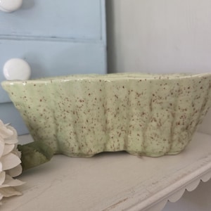 May include: A light green ceramic dish with a speckled pattern. The dish has a scalloped edge and a rectangular shape. It is sitting on a white shelf. A blue cabinet is in the background.