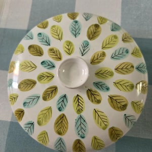 May include: A white ceramic lid with a central knob, decorated with a pattern of green, yellow, and teal leaf shapes. The leaves are arranged in a radial pattern, creating a decorative design. The lid is likely for a serving dish or container.