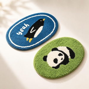 Peut inclure: Deux tapis ovales touffetés à la surface texturée. Un tapis est bleu avec une bordure blanche, représentant un manchot noir et blanc et le mot "yeah" en texte blanc. Le second tapis est vert et présente un motif de panda noir et blanc.