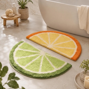 May include: Two semi-circular bath mats featuring a citrus fruit design. One mat is yellow and orange, resembling a lemon or orange slice, while the other is light and dark green, resembling a lime slice, both with white segment detailing.