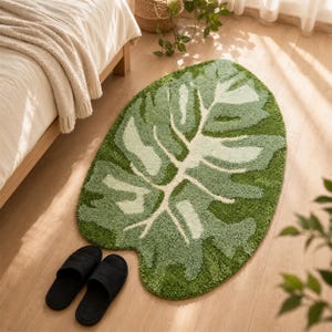 Peut inclure: Un tapis touffeté en forme de grande feuille de monstera, présentant un poil texturé dans diverses nuances de vert, incluant le vert forêt foncé, le sauge et le menthe clair, avec un motif de nervure centrale et une paire de chaussons noirs.