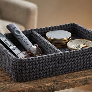 May include: A rectangular dark gray crocheted storage basket with two internal compartments. One side holds two black remote controls and a pair of eyeglasses. The other side contains a stack of round coasters and a small dish with metal keys.