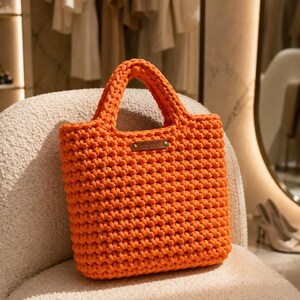 May include: A bright orange crochet tote bag featuring a textured, knotted stitch pattern and two integrated handles. A small, rectangular brown leather-like tag is attached to the front center, with the word HANDMADE embossed in gold lettering.