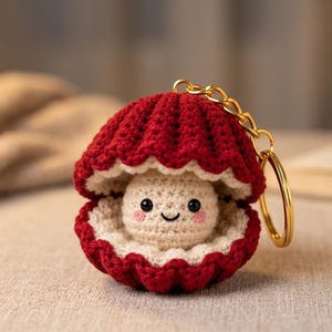 Puede incluir: Un llavero de ganchillo con forma de concha de color rojo oscuro que se abre para revelar una pequeña perla de color crema con una cara sonriente, ojos de cuentas negras y mejillas sonrosadas. El accesorio incluye una anilla y cadena doradas.