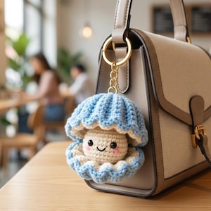 以下が含まれることがあります： ゴールドの留め具が付いたベージュの構造的なハンドバッグ。ハンドルには、かぎ針編みの水色の貝殻のキーホルダーが取り付けられています。キーホルダーの中には、笑顔が刺繍された小さなクリーム色のかぎ針編みの真珠が入っています。