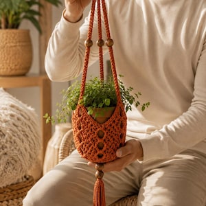 Puede incluir: Un colgador de plantas de macramé color naranja quemado con un diseño de cuerdas anudadas, tres cuentas de madera oscura integradas en los cordones superiores y una larga borla decorativa en la parte inferior, sosteniendo una maceta de terracota.