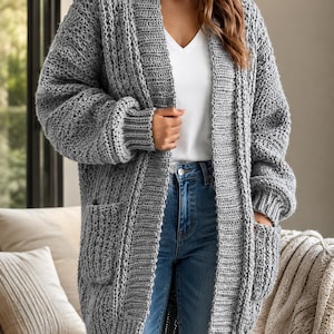 May include: A chunky knit grey cardigan featuring a ribbed shawl collar, long sleeves with ribbed cuffs, and two front patch pockets. The open-front garment is worn over a white v-neck top and blue denim jeans, showcasing a thick, textured yarn construction.