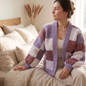 May include: A chunky crochet cardigan featuring a patchwork design of large squares in shades of lavender, mauve, and white. The garment has long sleeves with ribbed cuffs, a ribbed collar, and an open front, worn over a white top and beige trousers.