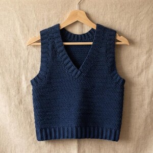 May include: A dark navy blue sleeveless sweater vest featuring a deep V-neckline and a ribbed hem. The garment is constructed with a visible crochet stitch pattern and is displayed on a wooden hanger, showcasing a solid color and textured knit.