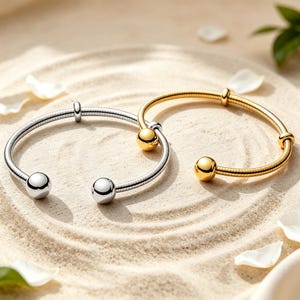Pode incluir: Duas pulseiras abertas, uma em tom prateado polido e outra em tom dourado polido. Cada pulseira apresenta um design de banda flexível e canelada com uma esfera lisa em cada extremidade e uma conta deslizante ao longo da banda.