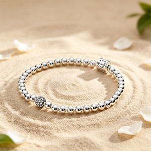 Könnte beinhalten: Ein silbernes Perlenarmband mit polierten, kugelförmigen Perlen, einer einzelnen texturierten Perle mit pavé-artiger Oberfläche und einem rechteckigen Silberverschluss mit der Gravur PANDORA. Das Armband ist kreisförmig angeordnet.