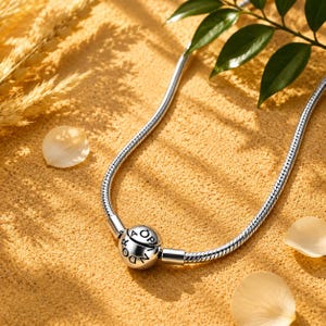May include: A silver snake chain necklace featuring a round clasp bead engraved with the text PANDORA. The chain has a smooth, flexible texture and a polished metallic finish, with the circular clasp serving as the central focal point.
