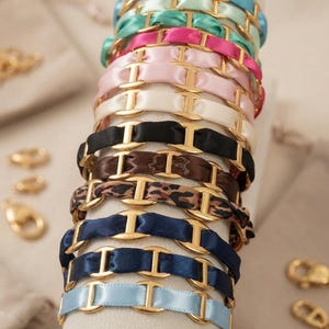 Peut inclure: Une collection de bracelets composés de maillons de chaîne en métal doré entrelacés avec des rubans de satin de différentes couleurs, notamment bleu clair, vert menthe, rose vif, rose pâle, crème, noir, marron, imprimé léopard et bleu marine.