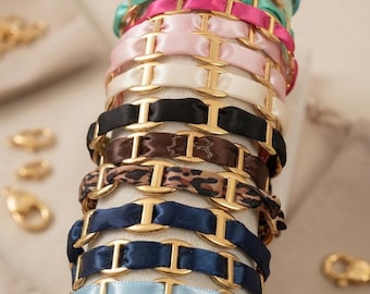 Può includere: Una collezione di bracciali con maglie metalliche dorate intrecciate con nastri di raso di vari colori. La pila include tonalità di azzurro, verde menta, turchese, rosa acceso, rosa pallido, crema, nero, marrone, stampa leopardata e blu navy.