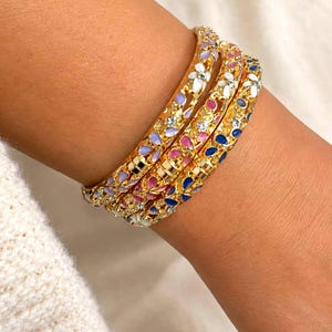 Peut inclure: Trois bracelets joncs en métal doré avec un motif floral ajouré et des pétales en émail incrustés dans des tons blanc, lavande, rose et bleu, agrémentés de petits cristaux transparents incrustés dans le métal.