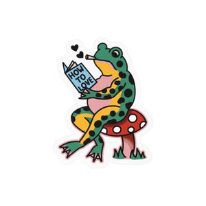 Peut inclure: Un autocollant découpé représentant une grenouille verte à taches noires assise sur un champignon rouge à pois blancs. La grenouille a un ventre rose, tient un livre bleu intitulé HOW TO LOVE, une cigarette à la bouche et deux cœurs noirs.