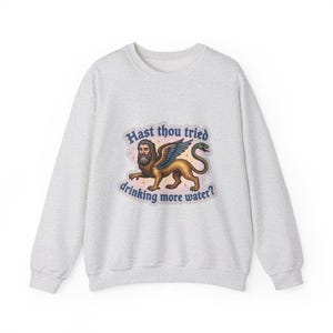 May include: A light grey crewneck sweatshirt featuring a central graphic of a winged lion with a human face and a snake tail. The text "Hast thou tried drinking more water?" is printed in dark blue gothic lettering around the illustration.
